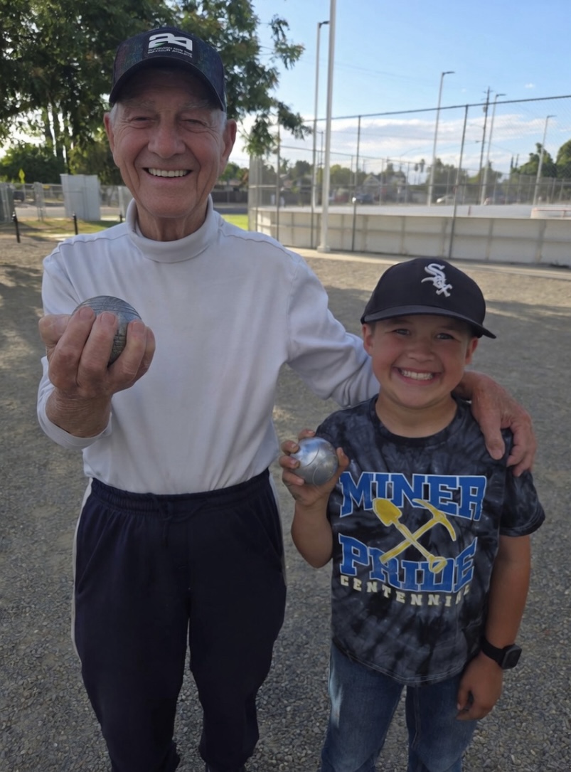 An older Fresno pétanque player sharing the game with a young player, both smiling and holding boules